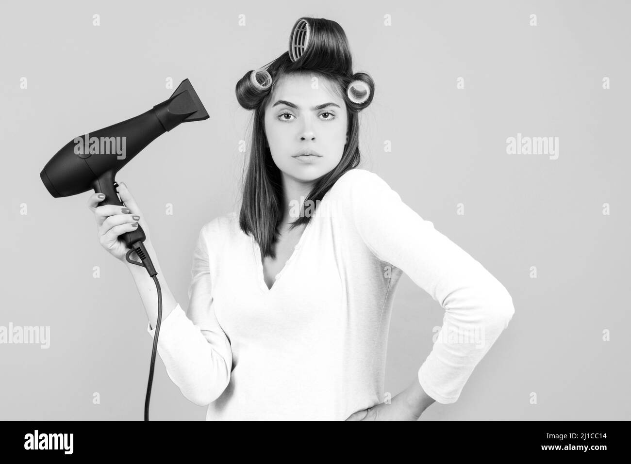 Young funny woman with hair dryer and rollers. Woman with hair dryer. Beautiful girl with straight hair drying hair with professional hairdryer. Stock Photo