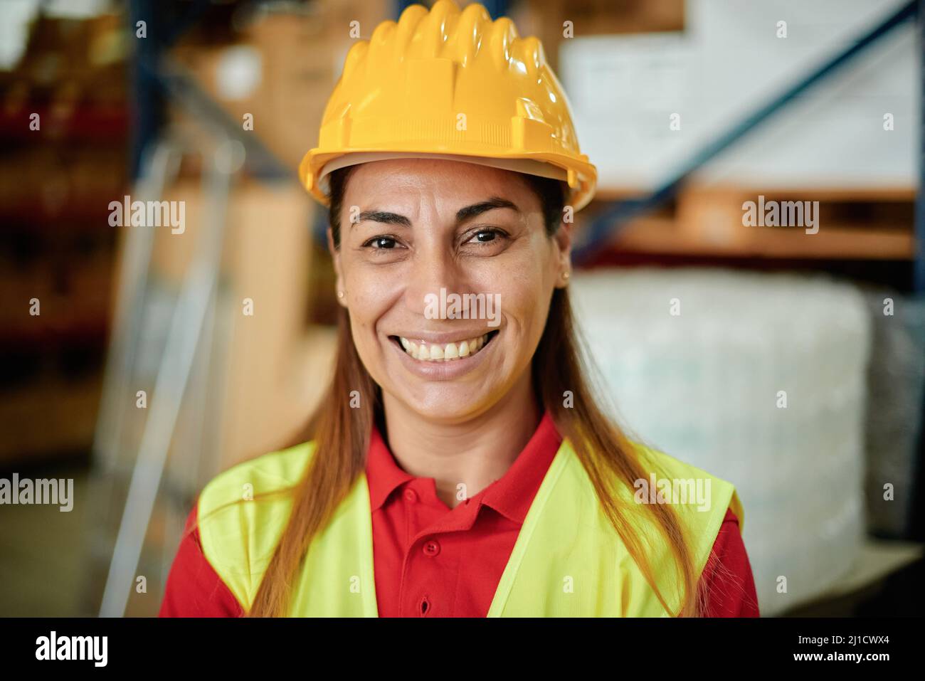 Portrait of a happy adult Caucasian working woman looking at the camera inside a warehouse wearing a hard hat and safety clothing - Focus on the Face Stock Photo