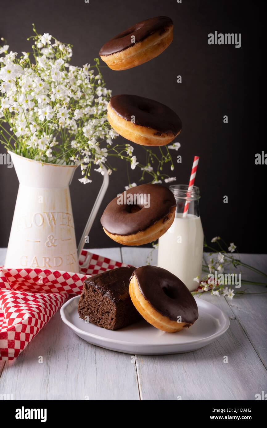 Homemade doughnuts covered with chocolate glaze falling on white plate and glass of milk on white rustic wooden surface. Levitation photography Stock Photo