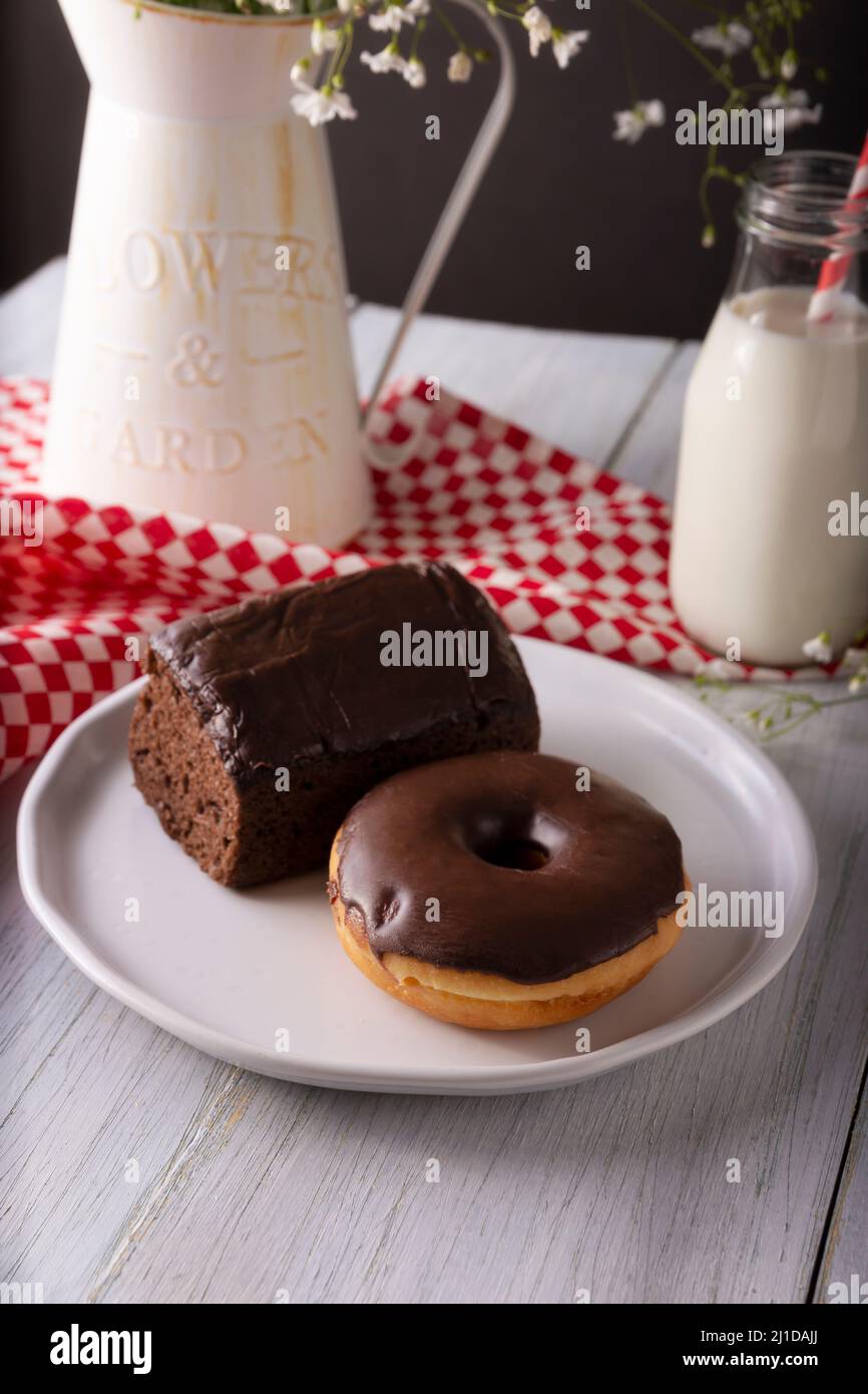 homemade chocolate donut and sponge cake covered with chocolate glaze and glass of milk on white rustic wooden surface Stock Photo