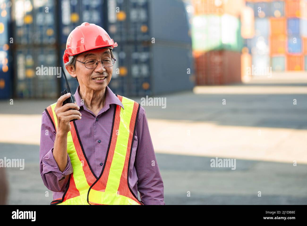 A senior elderly Asian worker engineer wearing safety vest and helmet standing and holding radio walkies talkie at logistic shipping cargo containers Stock Photo