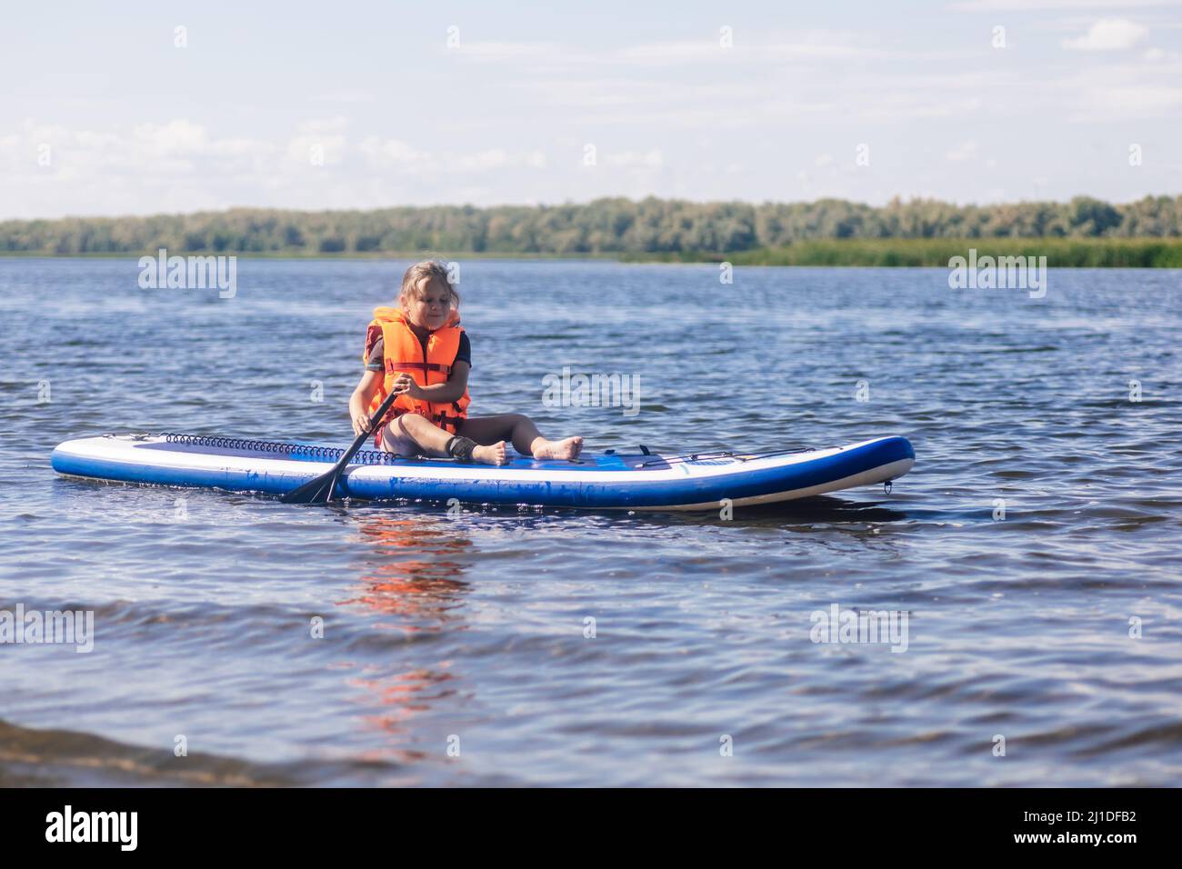Little blonde girl puddle boarding on lovely lake rowing with oar in hands looking at rippled water in vest life jacket. Active holidays. Inculcation Stock Photo