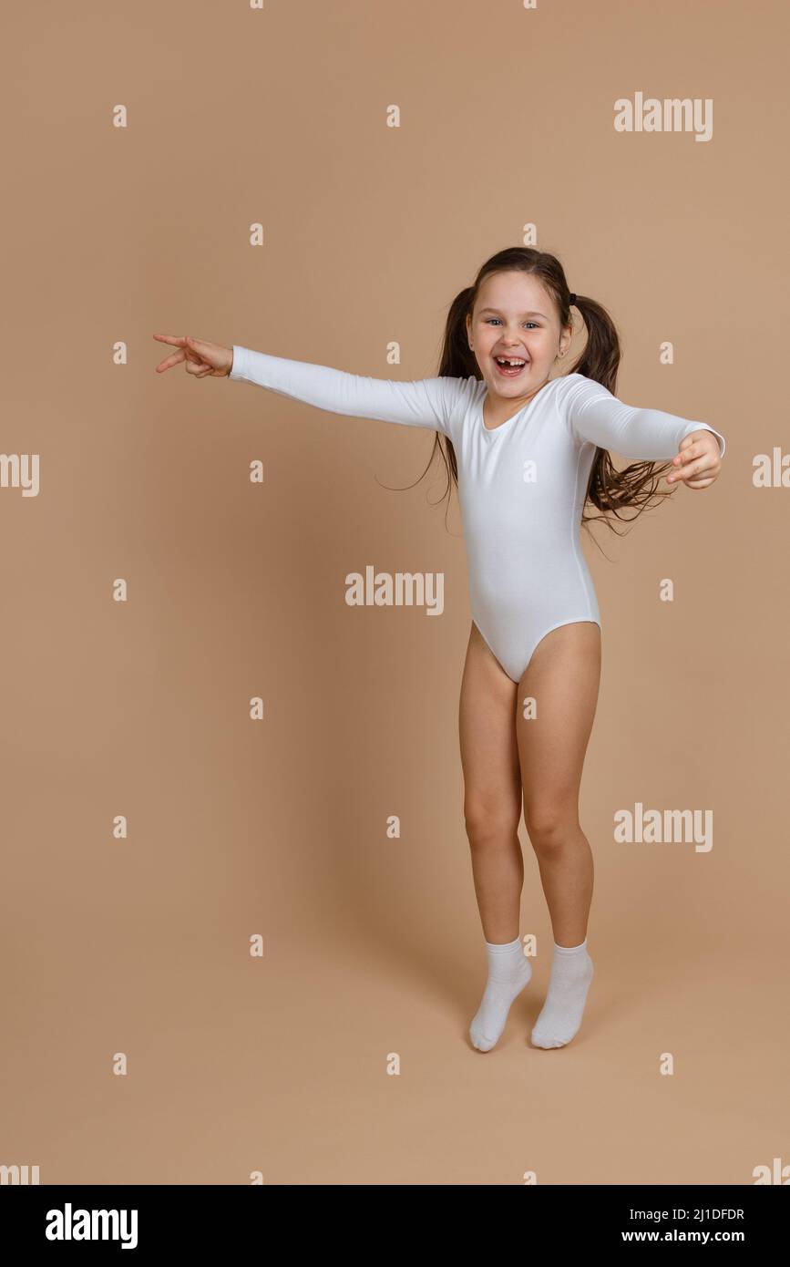 Portrait of young cute happy smiling girl with long dark hair in white training swimsuit and socks jumping on brown background, spreading hands Stock Photo