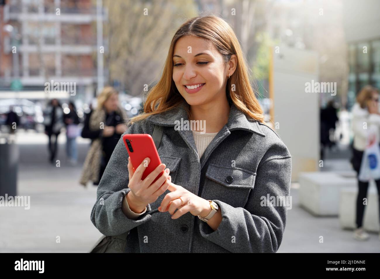 Pretty busy girl looking at mobile screen and smiling outside with blurred people on the background Stock Photo