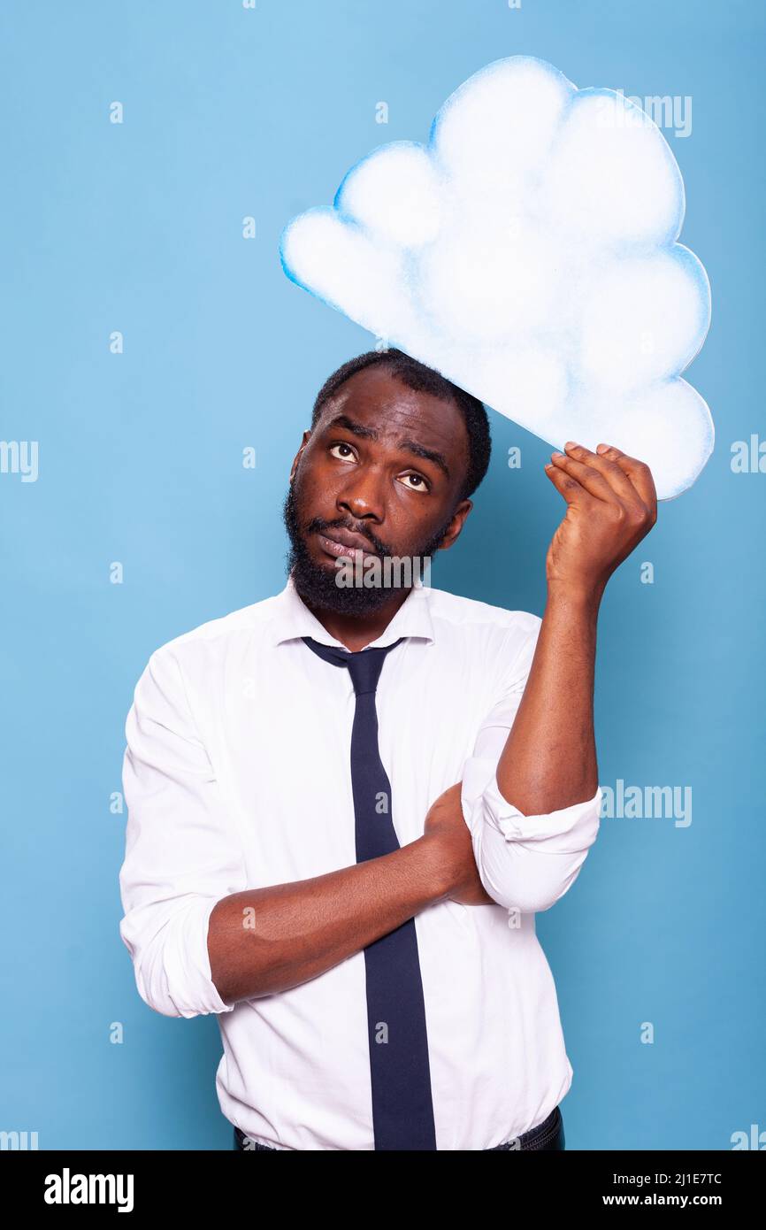 Portratit of accountant in white shirt looking up at white paper idea cloud brainstorming standing in front of blue background. African american entrepreneur thinking under thought bubble. Stock Photo