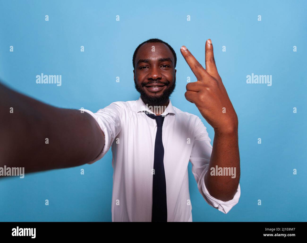 Smiling vlogger showing peace sign at camera in videocall conference on blue background. Wide angle pov of happy influencer taking a smartphone selfie raising two fingers gesture for social media. Stock Photo