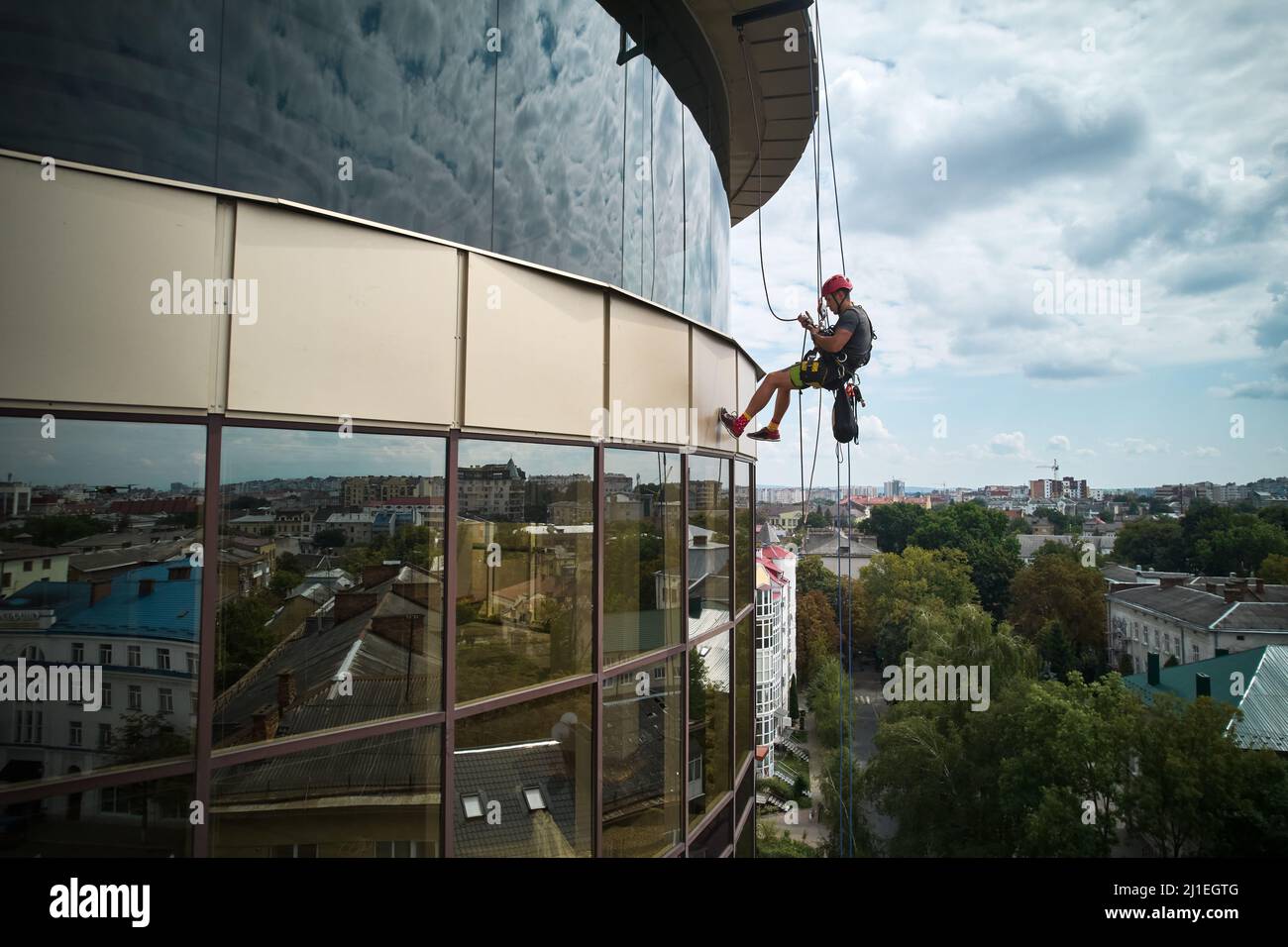 Industrial mountaineering worker cleaning glass window of high-rise building. Male cleaner using safety lifting equipment while washing window of skyscraper. Stock Photo