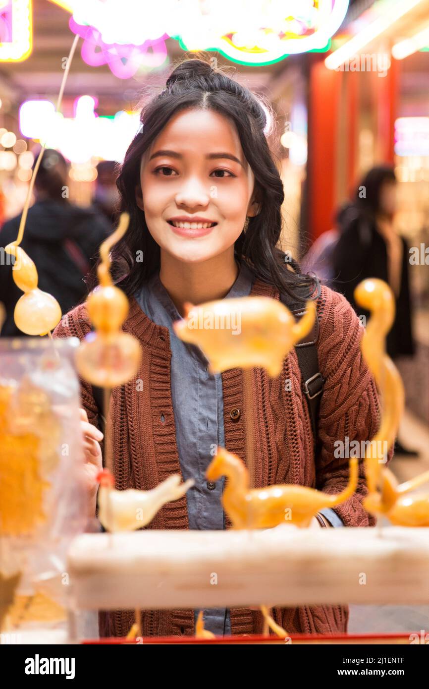 Young female college student buying and choosing calabash sugar figure at a night market in Beijing - stock photo Stock Photo