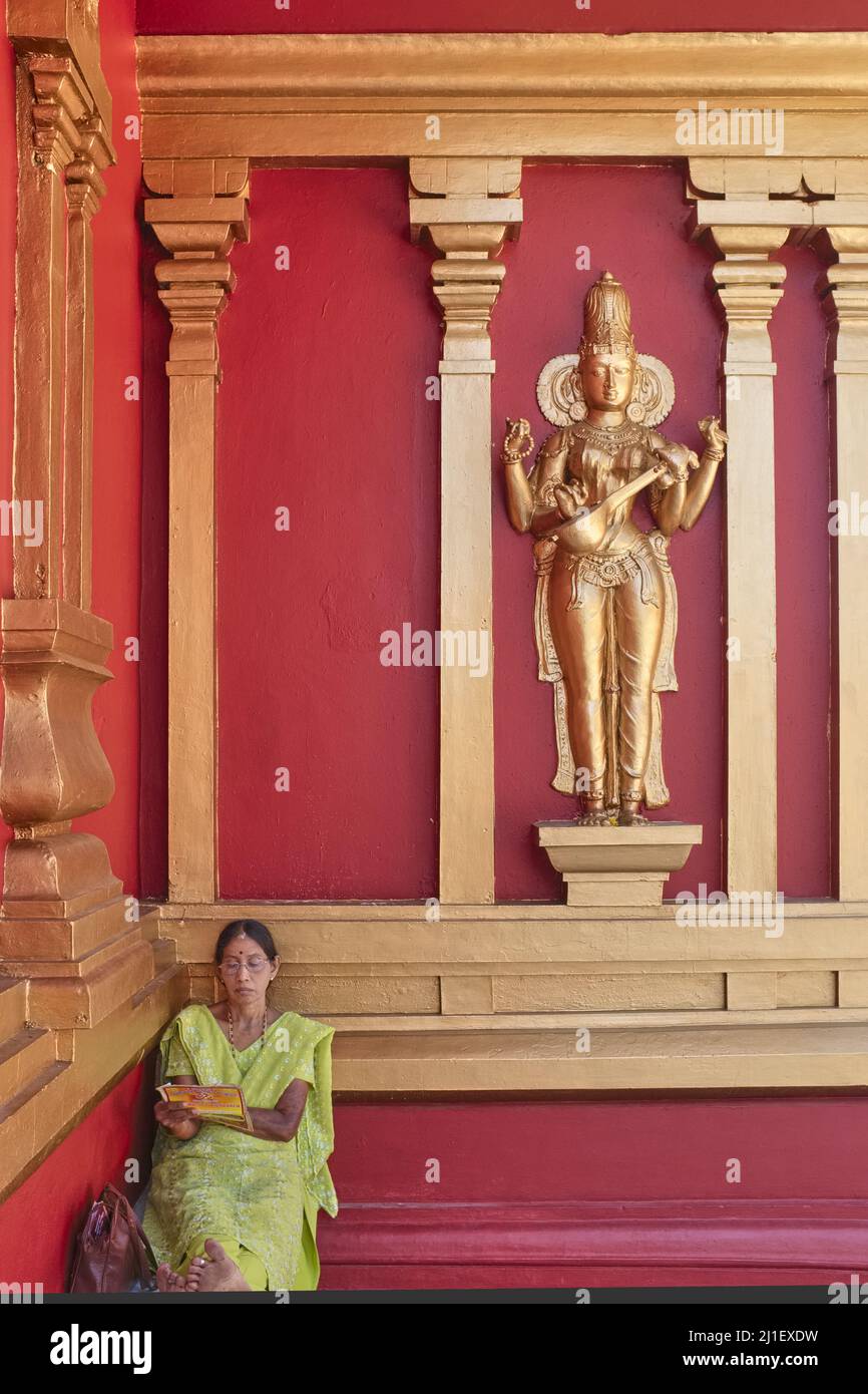 A woman sits by the wall of Kudroli Shree Gokarnanatheshwara Temple in Mangalore, Karnataka, India, next to a golden relief of Hindu goddess Saraswati Stock Photo