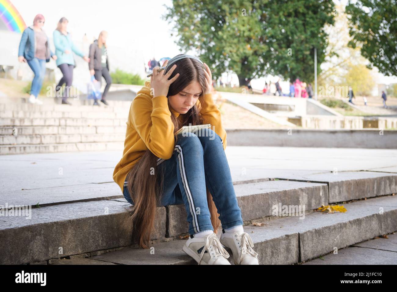 A sad teenage girl sits on the stairs and covers her head with her ...