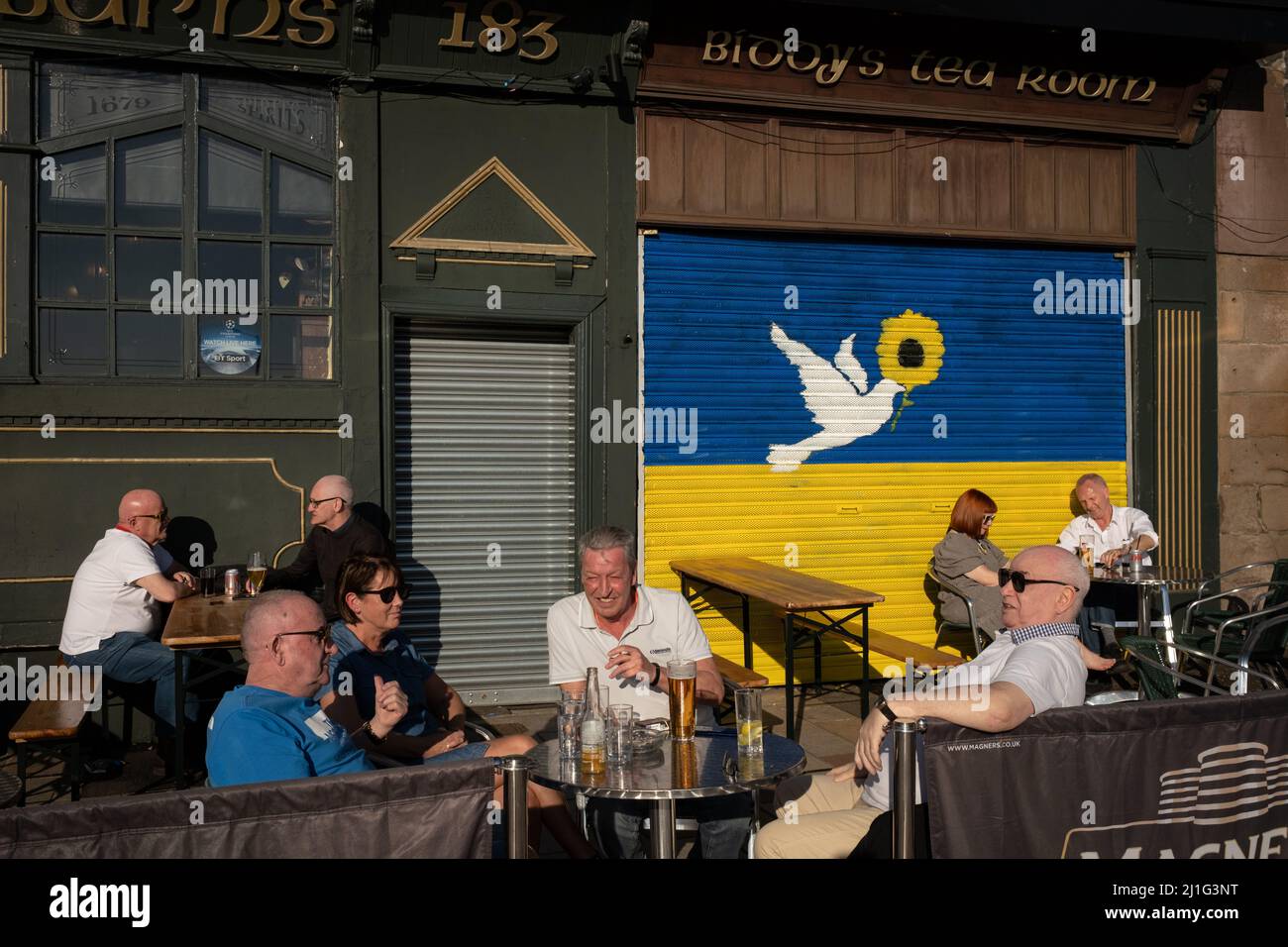 Glasgow, UK, 25th March 2022. People enjoy a drink in the sun in front of a mural on the wall of BiddyÕs Tea Room depicting the Ukrainian flag and a dove of peace, painted to show support for Ukraine in their ongoing war against the invasion by Russia, in the east end of Glasgow, Scotland, 25 March 2022. Photo credit: Jeremy Sutton-Hibbert/Alamy Live News. Stock Photo