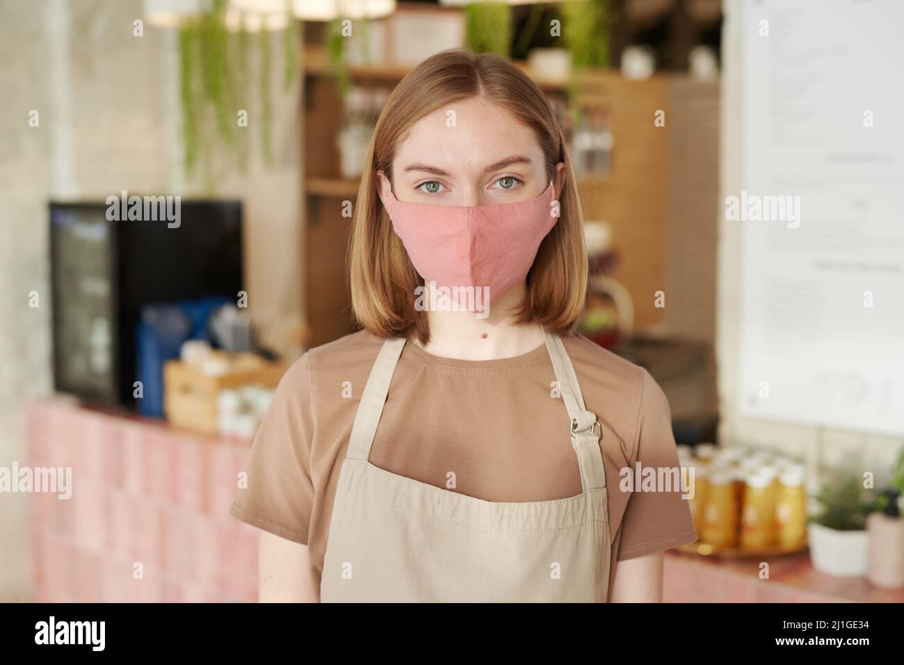 Horizontal medium close-up portrait of young woman wearing apron and protective mask starting workday in cafe looking at camera Stock Photo