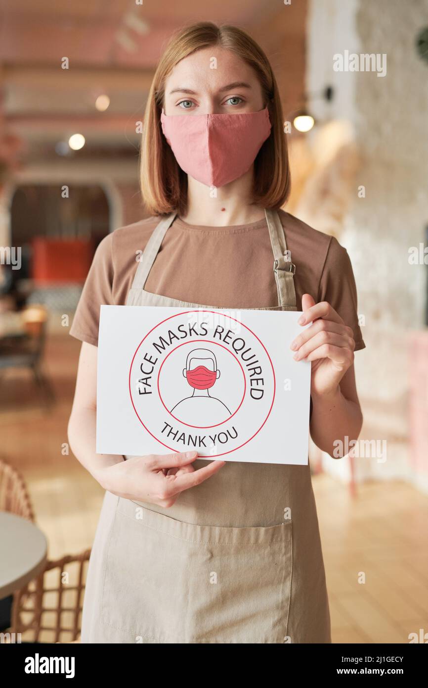 Vertical shot of unrecognizable female cafe worker standing alone holding Face Mask Required sign looking at camera Stock Photo