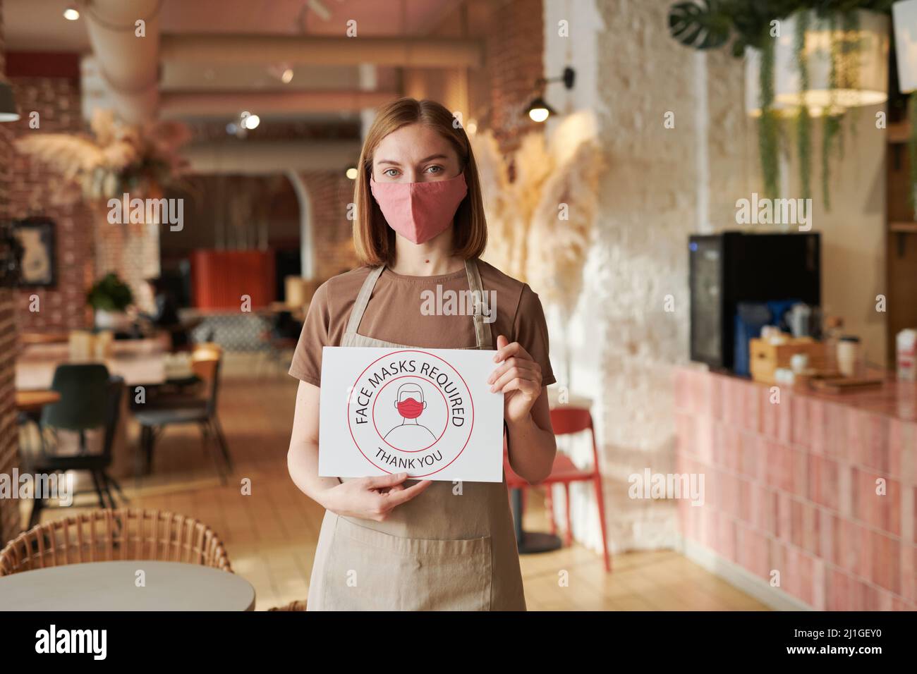 Horizontal medium shot of unrecognizable female waitperson holding Face Mask Required sign looking at camera Stock Photo