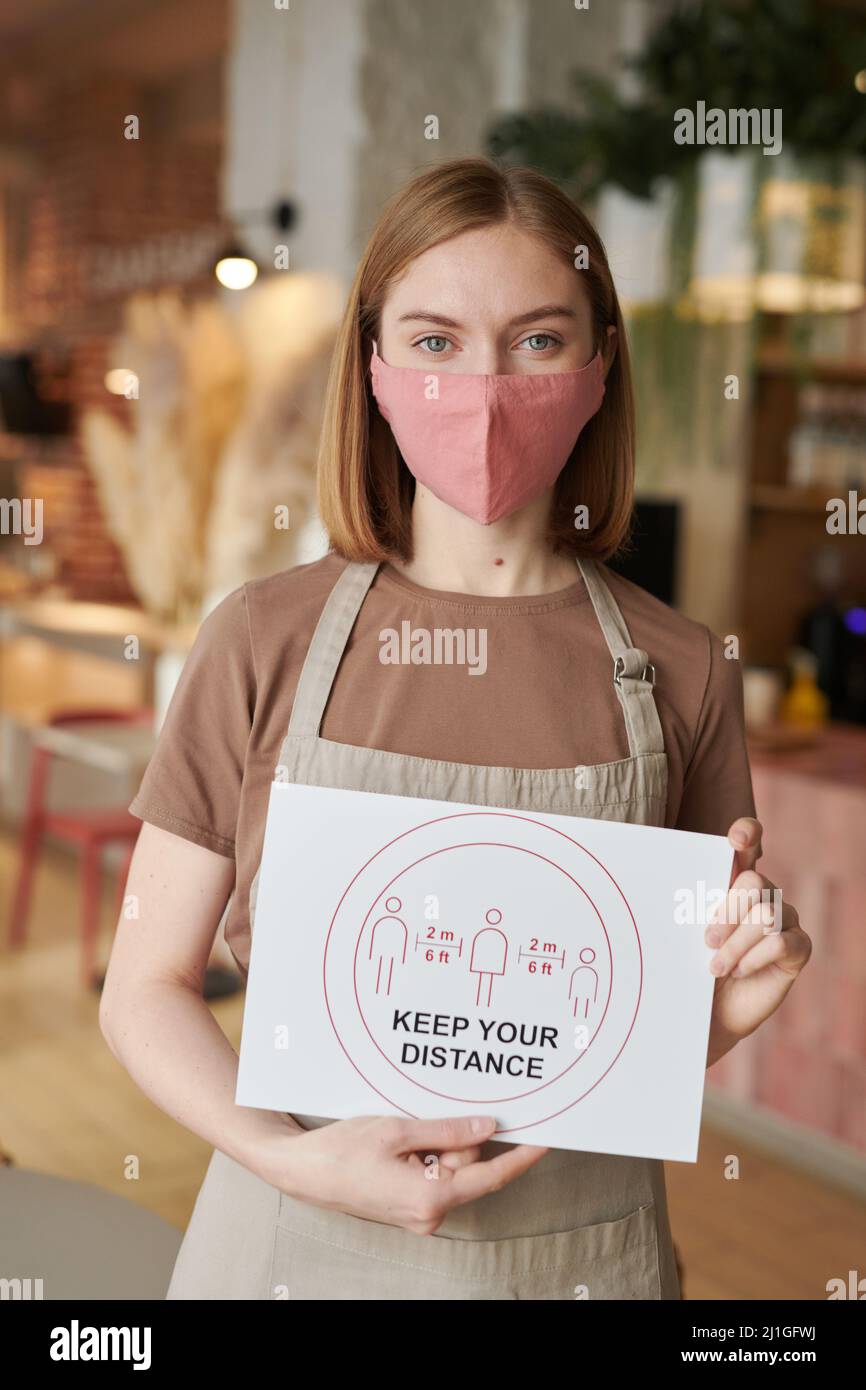 Vertical medium portrait shot of unrecognizable waitperson wearing apron standing in cafe holding Keep Your Distance sign looking at camera Stock Photo