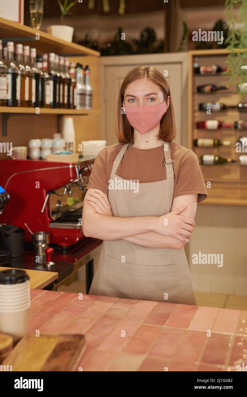 Vertical medium long shot of modern female waitperson wearing protective mask standing at counter with arms crossed looking at camera Stock Photo