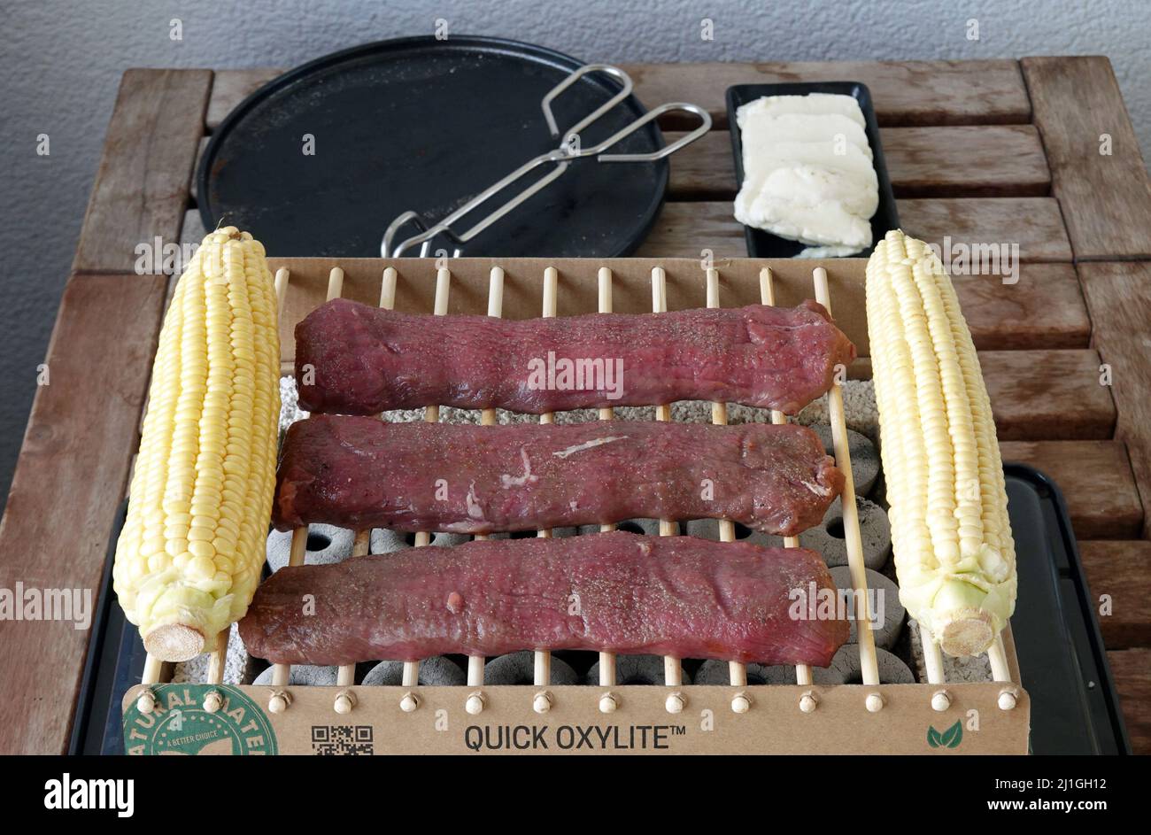 Berlin, Germany. 17th July, 2021. A disposable grill with the grate made of bamboo and lamb (lamb salmon) and corn on the cob on it, stands on a table after lighting the stones. Next to it is a plate with tongs. The Instandgrill is made of bamboo, wood and lava stones and is one hundred percent biodegradable, according to the manufacturer. According to the instruction leaflet, the CasusGrill is guaranteed to last for one hour. Credit: Soeren Stache/dpa-Zentralbild/ZB/dpa/Alamy Live News Stock Photo