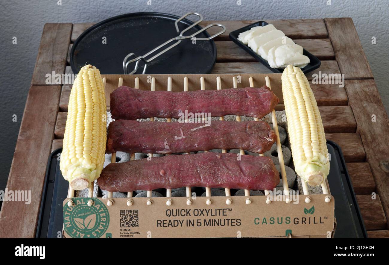 Berlin, Germany. 17th July, 2021. A disposable grill with the grate made of bamboo and lamb (lamb salmon) and corn on the cob on it, stands on a table after lighting the stones. Next to it is a plate with tongs. The Instandgrill is made of bamboo, wood and lava stones and is one hundred percent biodegradable, according to the manufacturer. According to the instruction leaflet, the CasusGrill is guaranteed to last for one hour. Credit: Soeren Stache/dpa-Zentralbild/ZB/dpa/Alamy Live News Stock Photo