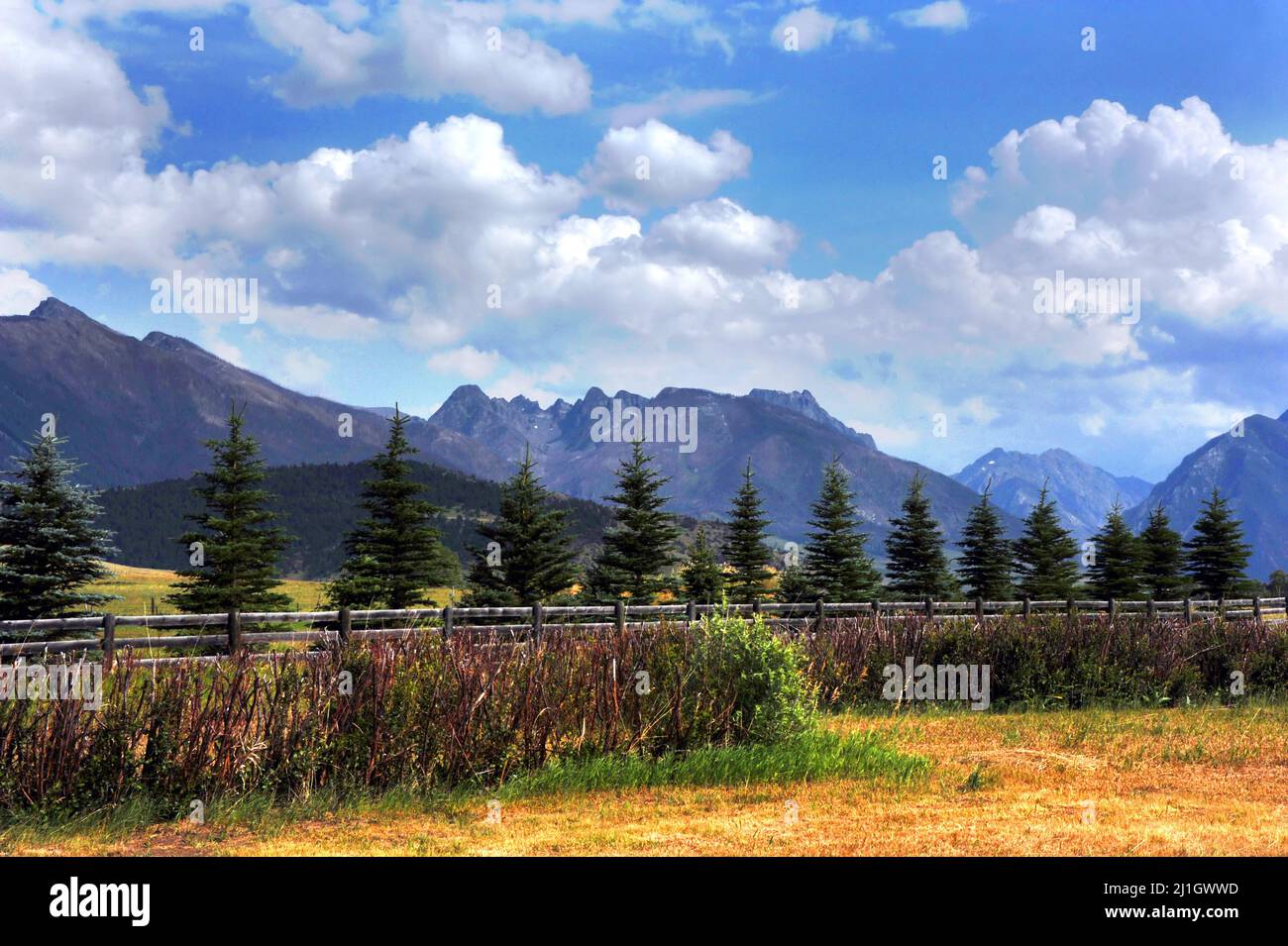 Rustic, wooden fence runs across photo, seperating field from distant, Absaroka mountains in Montana.  Blue sky and fluffy clouds complete image. Stock Photo