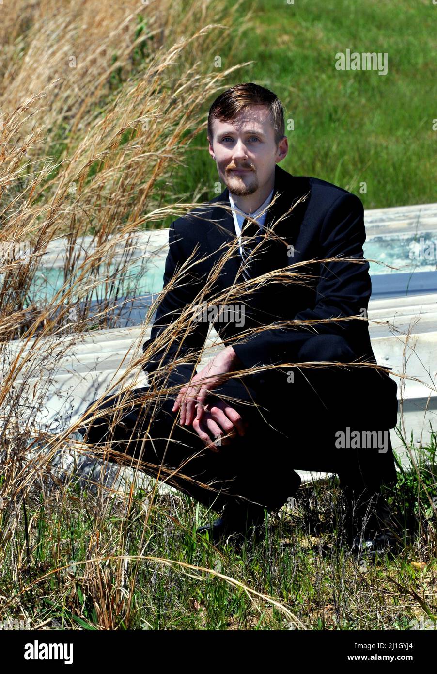 Rustic aluminum boats and golden grass surround handsome young man wearing a black business suit.  Sun is shining and man wears a small smile. Stock Photo