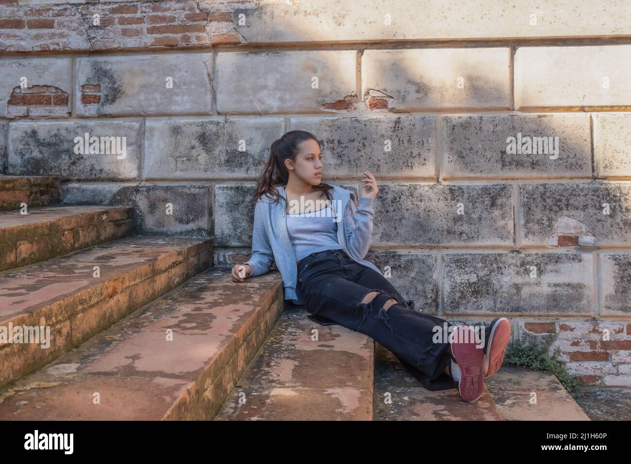 Young woman sits on grey stairs dressed in ripped jeans brick wall behind Stock Photo