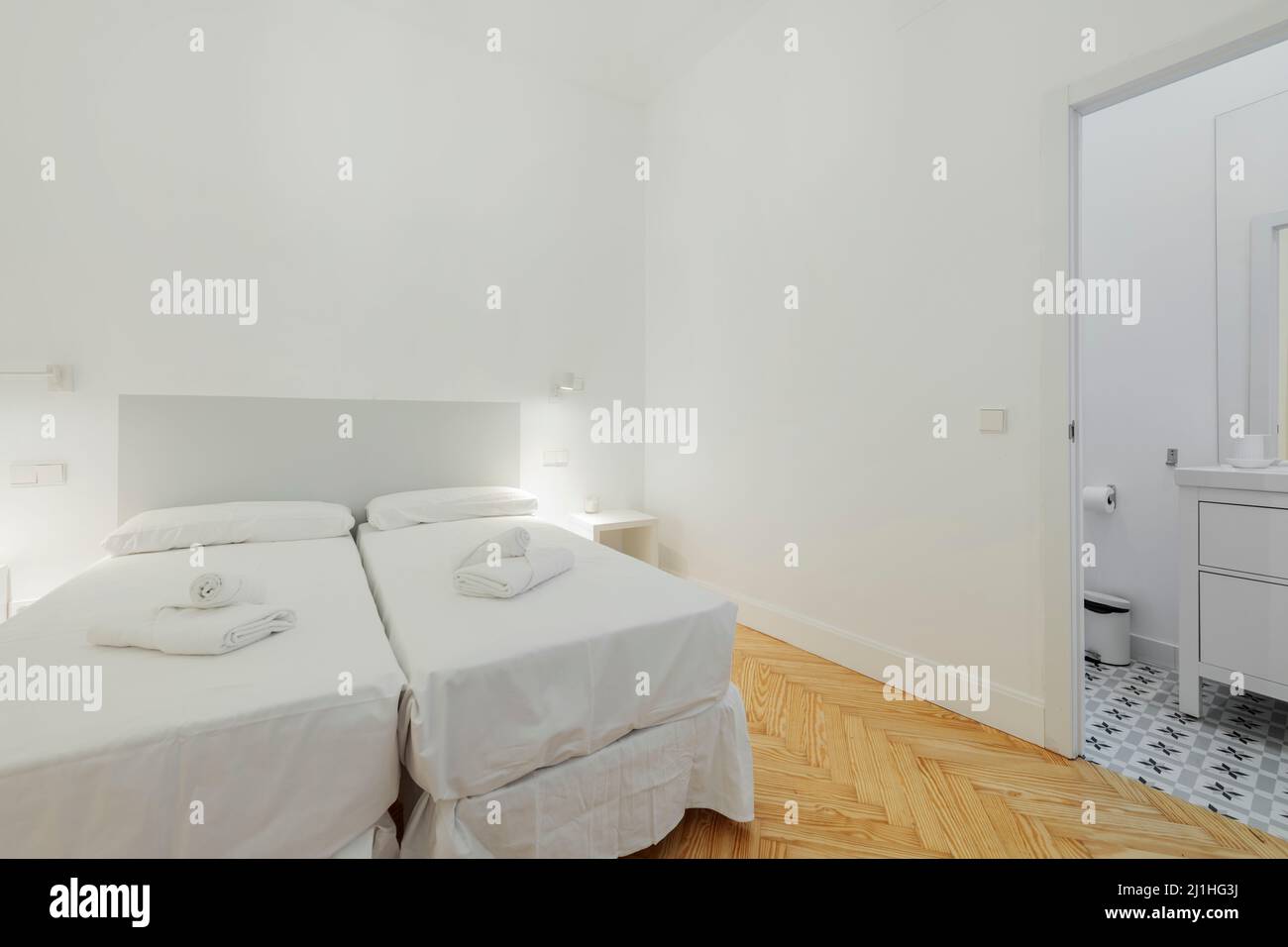 Bedroom with double beds and white sheets, bath towels on the beds and white bedside tables and access to an en-suite bathroom with white cabinets Stock Photo