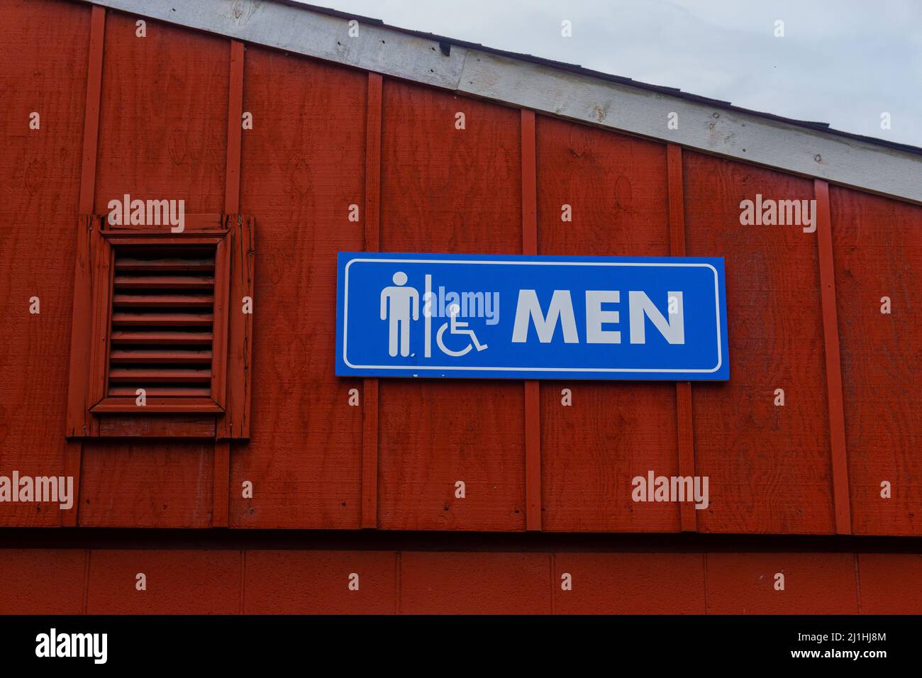 Men’s bathroom sign on the top of a building next to roof, colored blue and white in front of red wall. Stock Photo