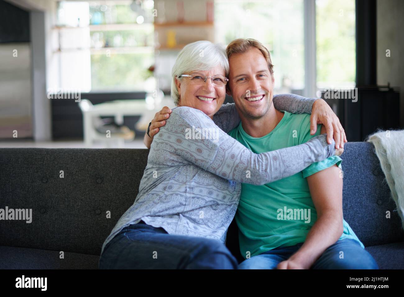 I love having him offer. Portrait of a senior woman embracing her son while sitting on the sofa at home. Stock Photo