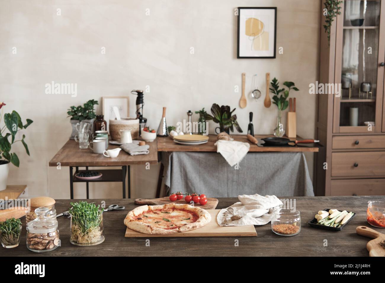 Prepared pizza on wooden board placed on table with ingredients and jars of spices, table with utensils and crockery in background Stock Photo