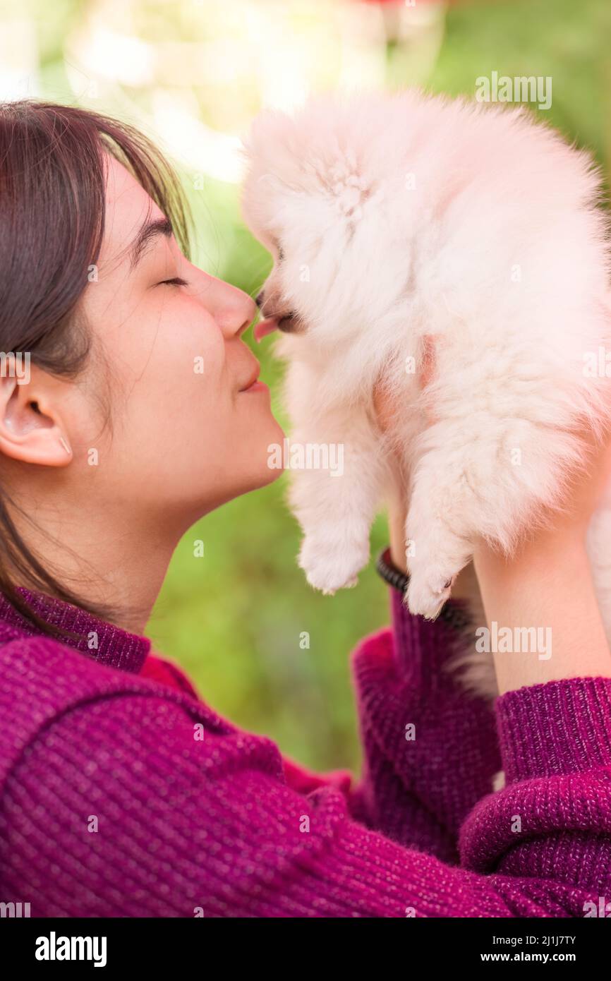 Biracial young woman in purple sweater holding small white pomeranian puppy outdoors on deck Stock Photo