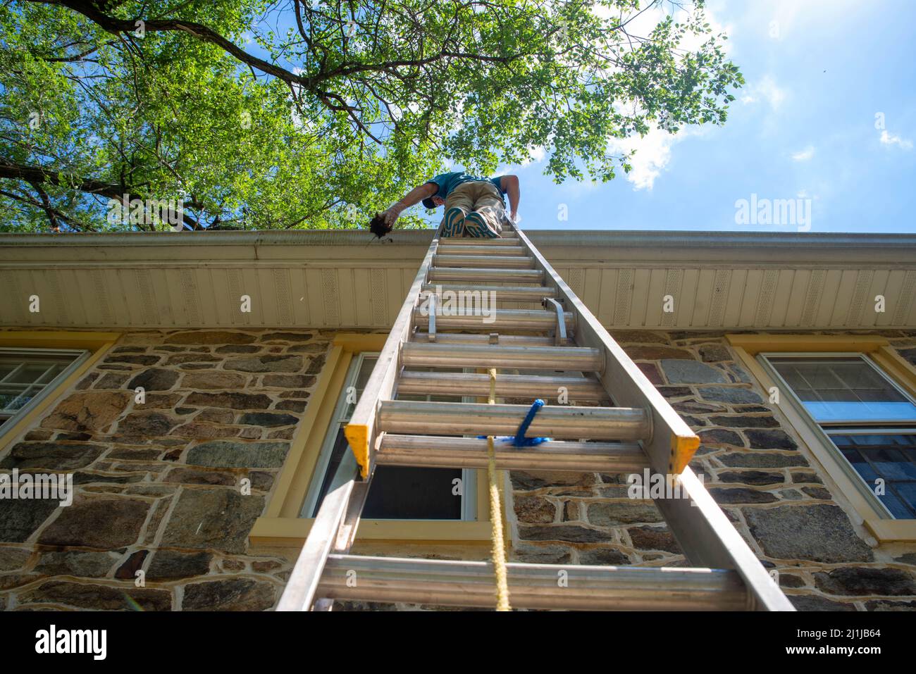 Low angle view of a man on a ladder cleaning gutters of old stone home Stock Photo