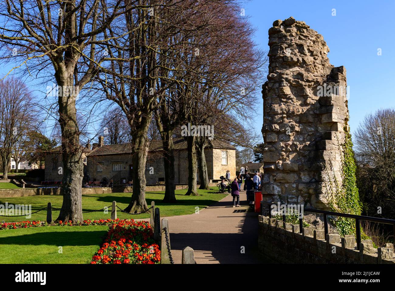 People relaxing, walking on paths in sunny park (bright border flowers, ancient ruins, blue sky) - Knaresborough Castle, North Yorkshire, England, UK. Stock Photo