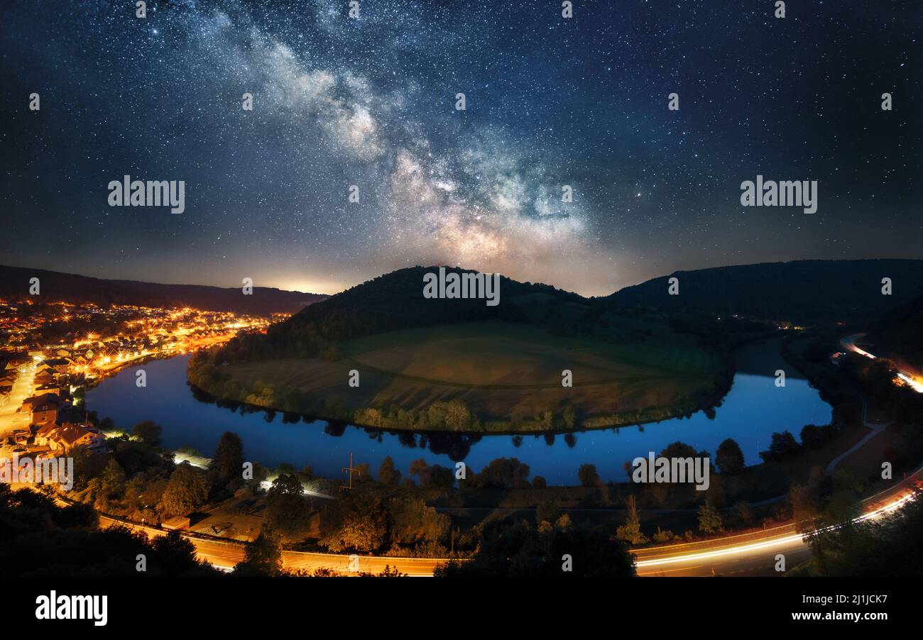 Majestic night sky with the Milky Way over a river bend around a hill, framed with an illuminated road and town Stock Photo