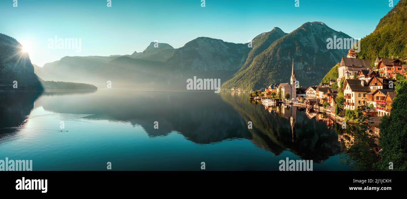 The iconic Austrian town Hallstatt in early morning light, a panoramic gorgeous landscape with the mountains, the houses and the teal sky reflected in Stock Photo