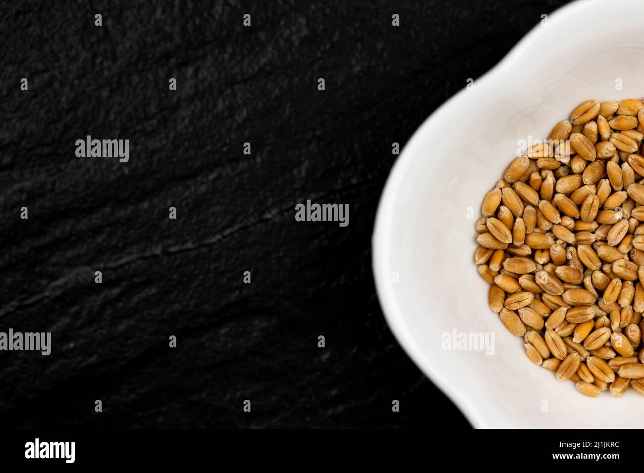 Top view of wheat grain in white bowl standing on black, stone surface. Scene lit with artificial, soft light. Stock Photo