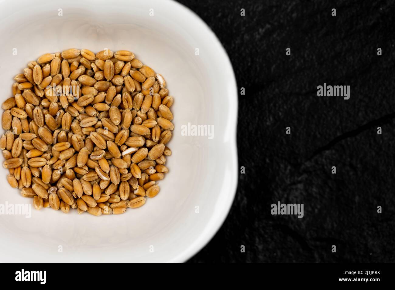 Top view of wheat grain in white bowl standing on black, stone surface. Scene lit with artificial, soft light. Stock Photo