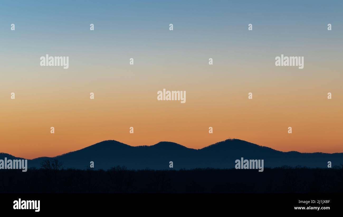 Silhouette of rolling hills surrounded with forest at twilight against blue sky with orange glow at horizon, landscape with mountain and woods at dusk Stock Photo