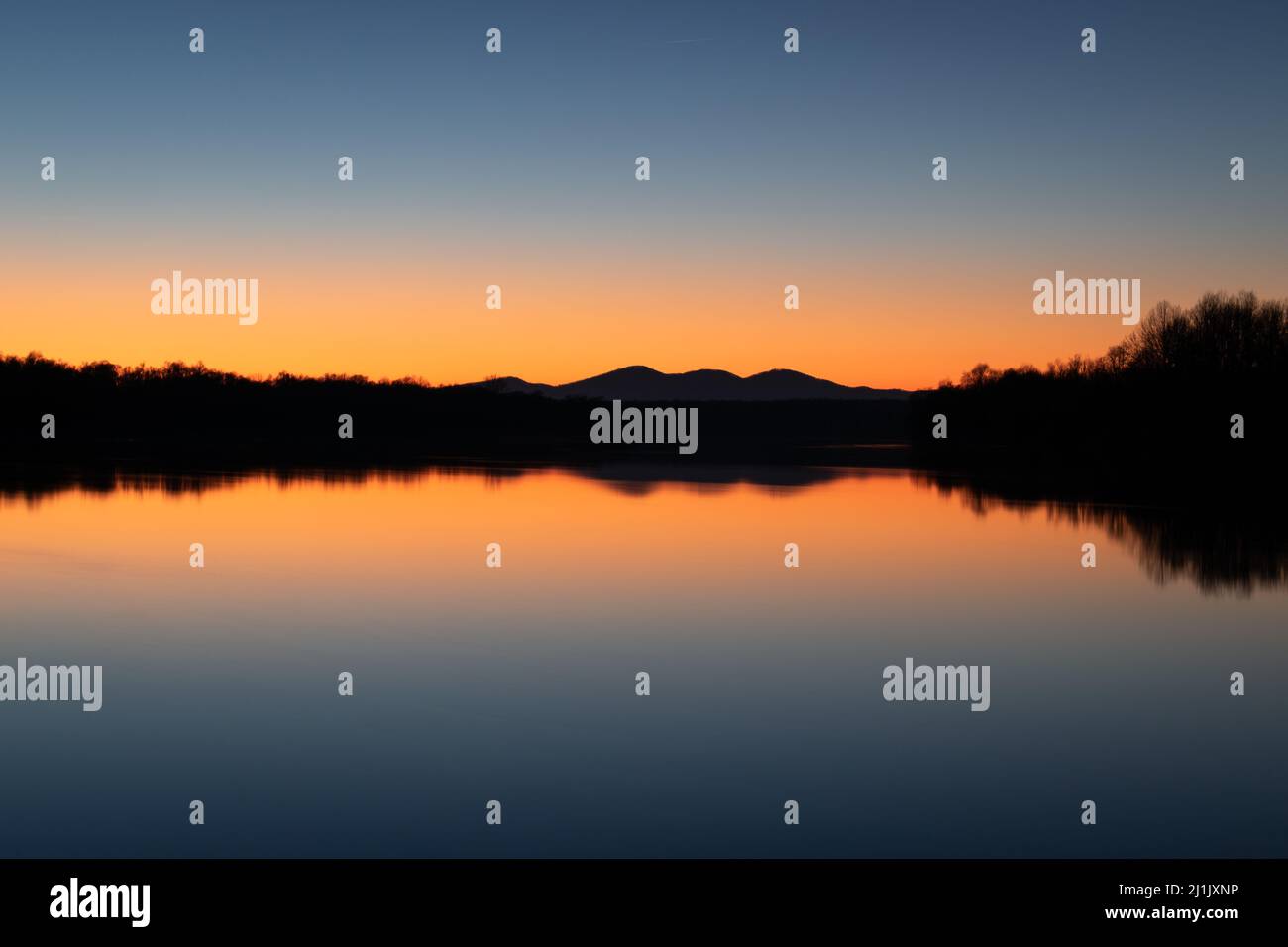 Landscape of silhouette of mountain and forest with reflection in river water against clear blue sky with orange glow at horizon at twilight Stock Photo