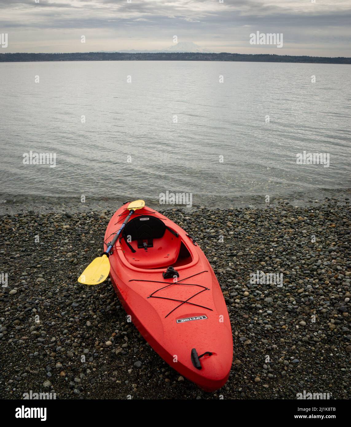 A bright red kayak with a yellow paddle on the rocky shore of Puget Sound Stock Photo