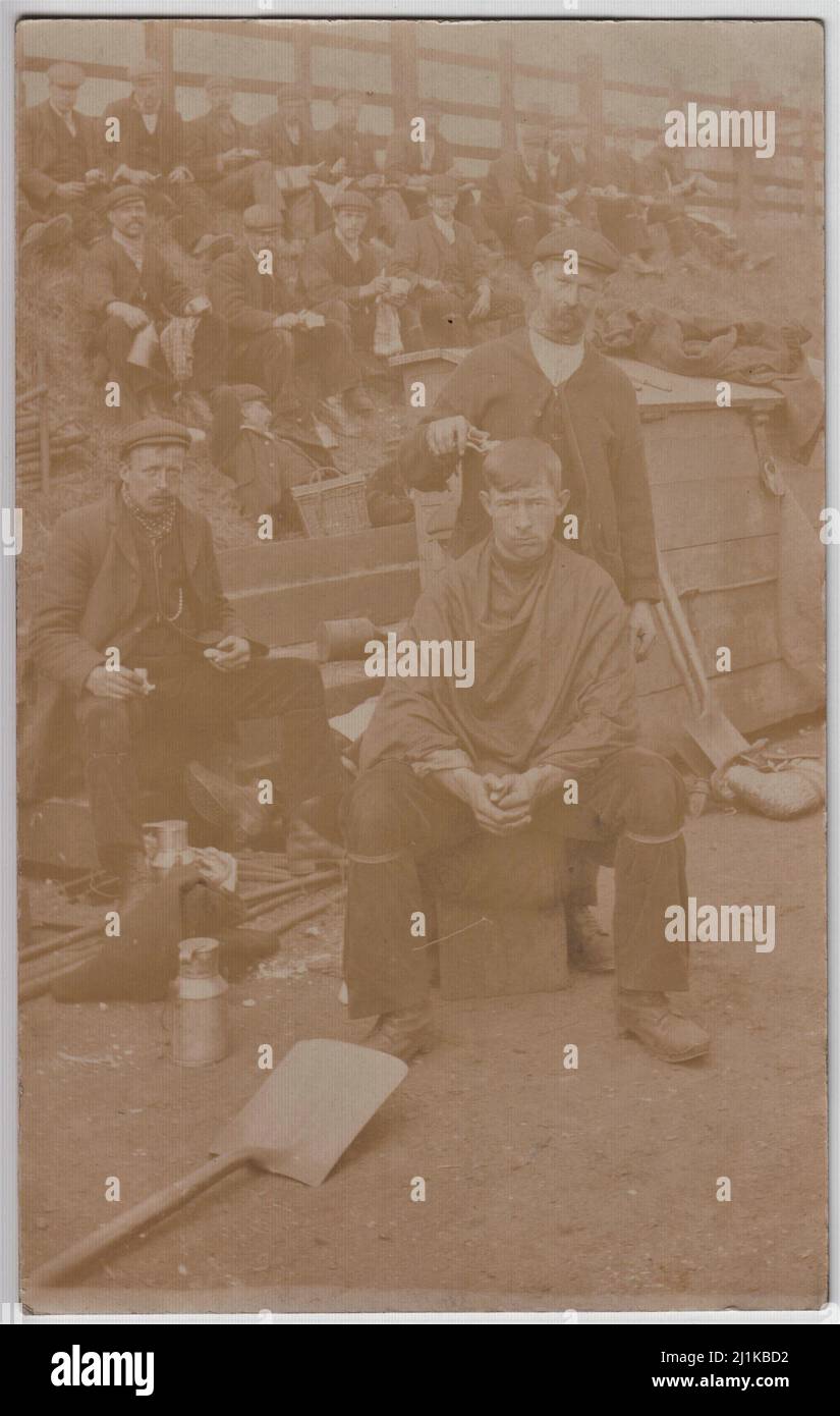Haircut, food & drink - navvies at rest, early 20th century. Group of men sitting on a bank having food and drink. One man is sitting in the foreground having his hair cut with a pair of clippers. Spades, metal flasks, flagons, a basket and other items are scattered around Stock Photo
