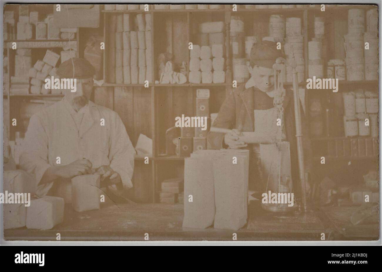 Behind the counter of an early 20th century grocers' shop. The grocer is packing up some goods whilst a young boy is using a set of scales. Various products can be seen on the shelves behind (one of the shelves has 'Sunlight Soap' written on it) Stock Photo