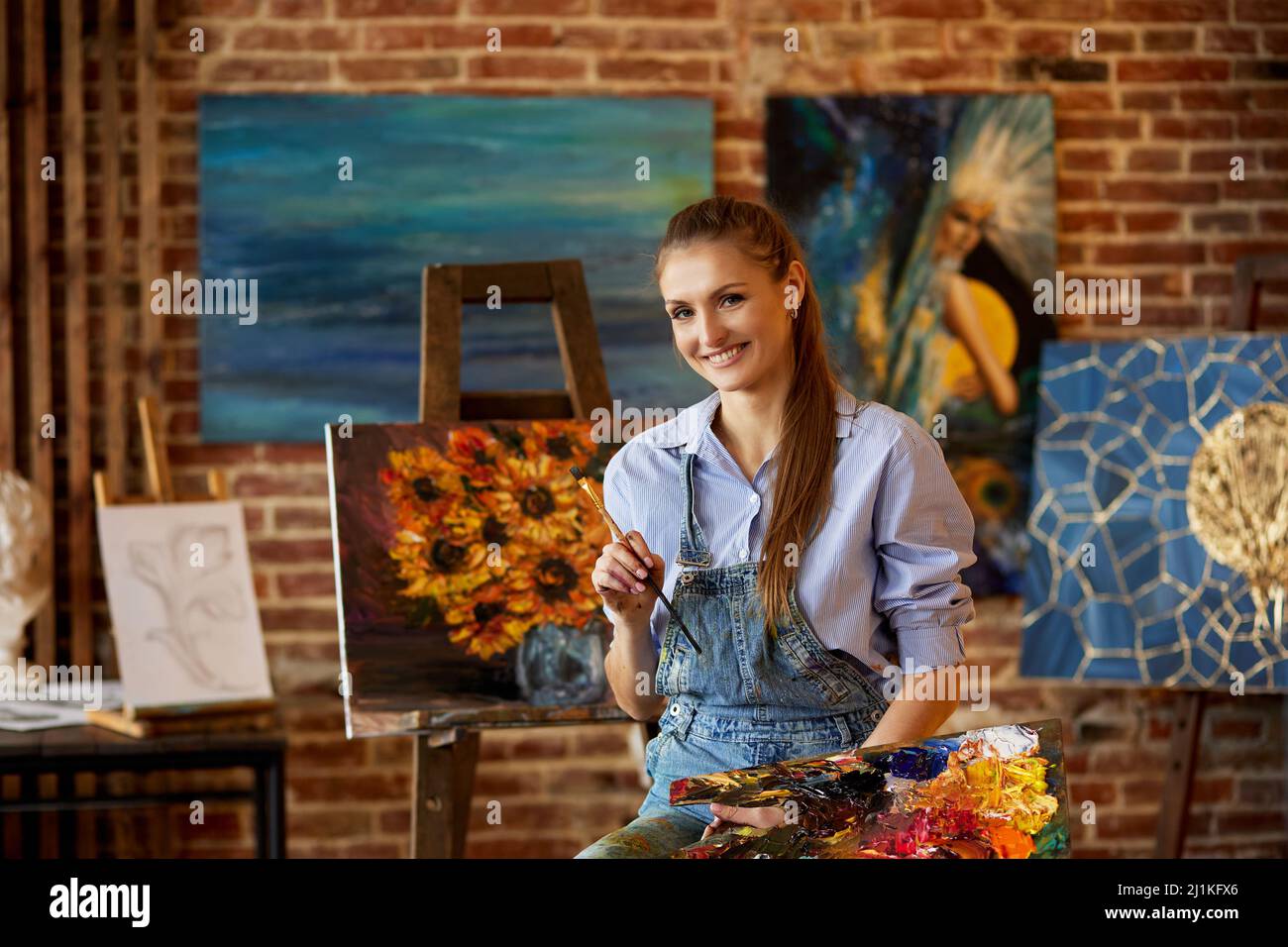 Portrait of young smiling female artist with her artworks in art studio ...