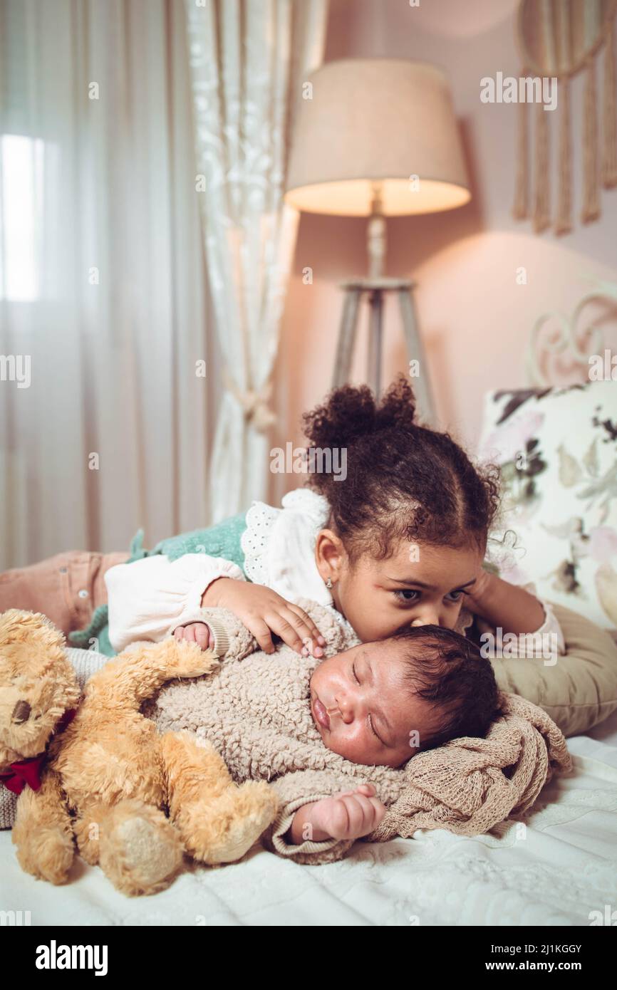 Little multiethnic girl, sister kissing her baby infant brother while they are lying on the bad Stock Photo