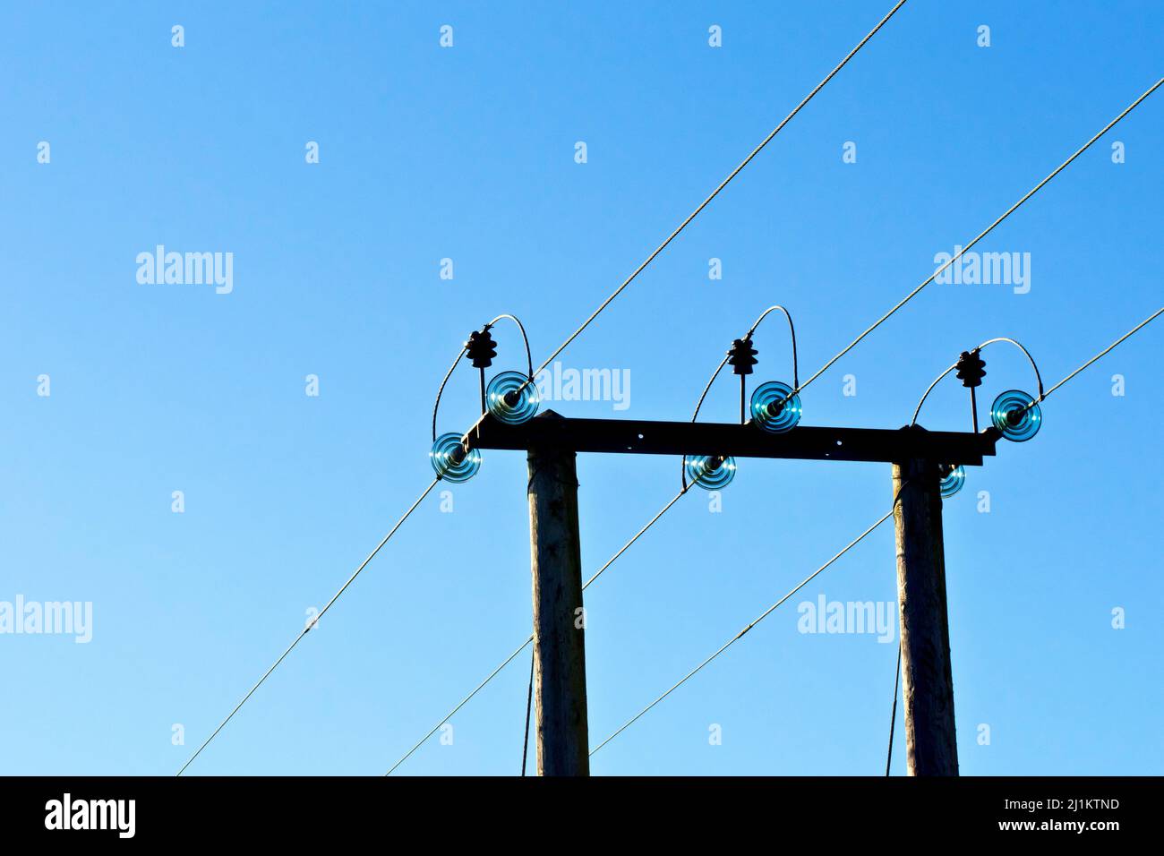 A rural wooden electricity pylon carrying three high voltage power lines across a field, shot back lit against a bright blue sky. Stock Photo
