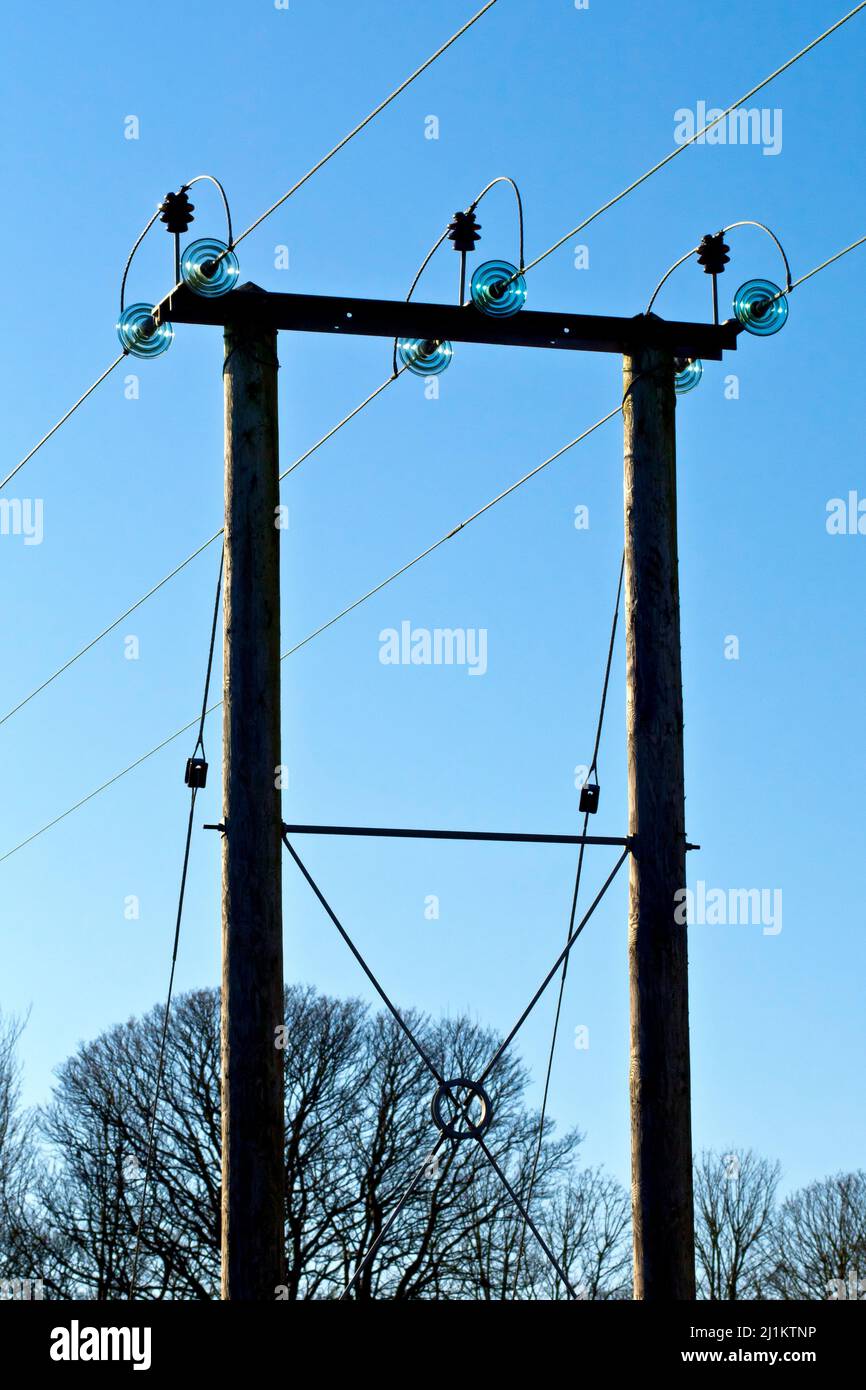 A rural wooden electricity pylon carrying three high voltage power lines across a field, shot back lit against a bright blue sky. Stock Photo