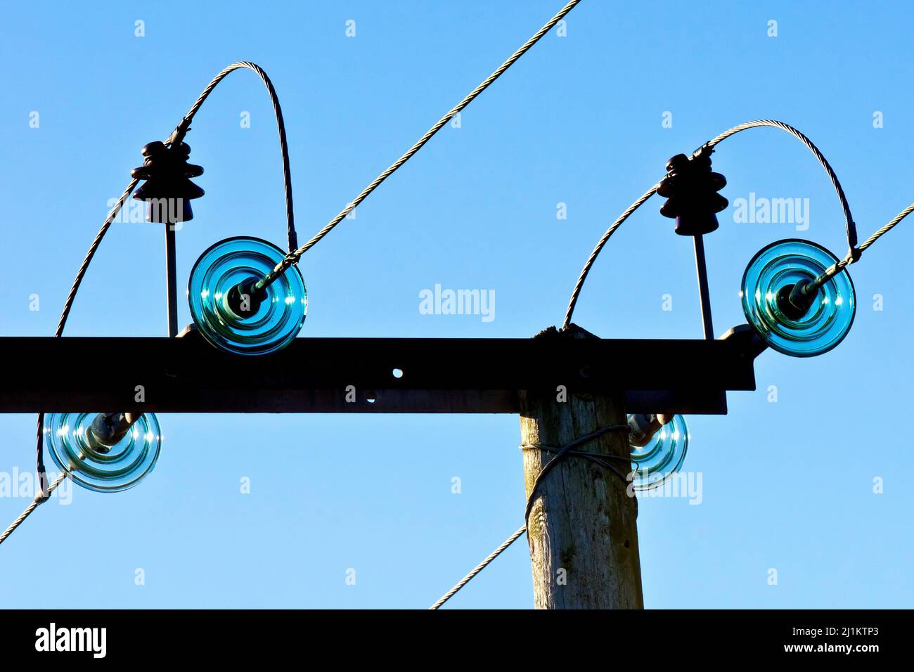 Close up of the top of a rural wooden electricity pylon showing the resistors and bridging cables, back-lit against a bright blue sky. Stock Photo