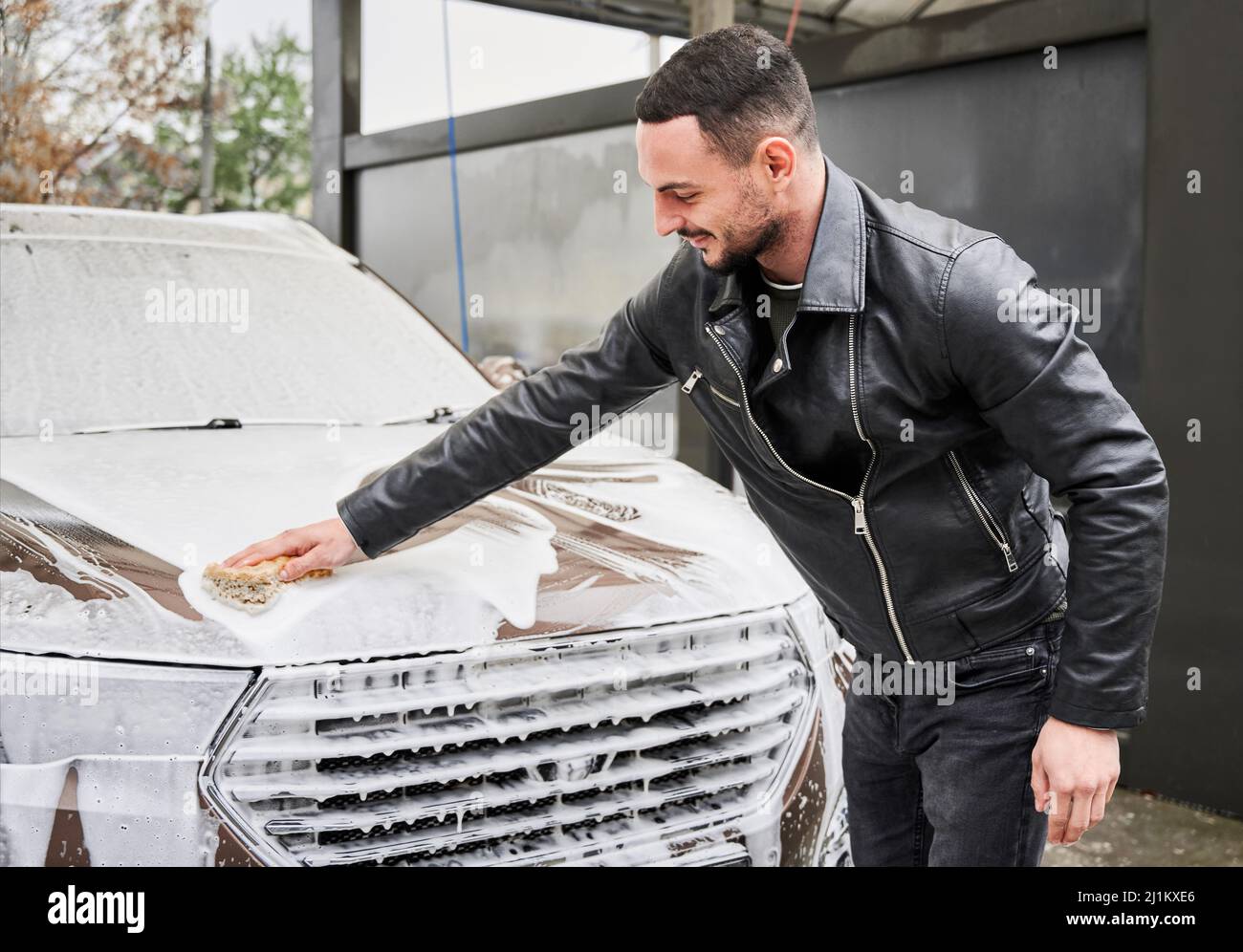 Smiling young man washing the hood of his soapy auto by hand with small
