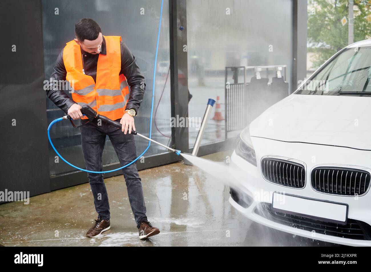 Young man washing car on carwash station outdoor, wearing orange vest. Handsome worker cleaning white automobile, using high pressure water. Stock Photo