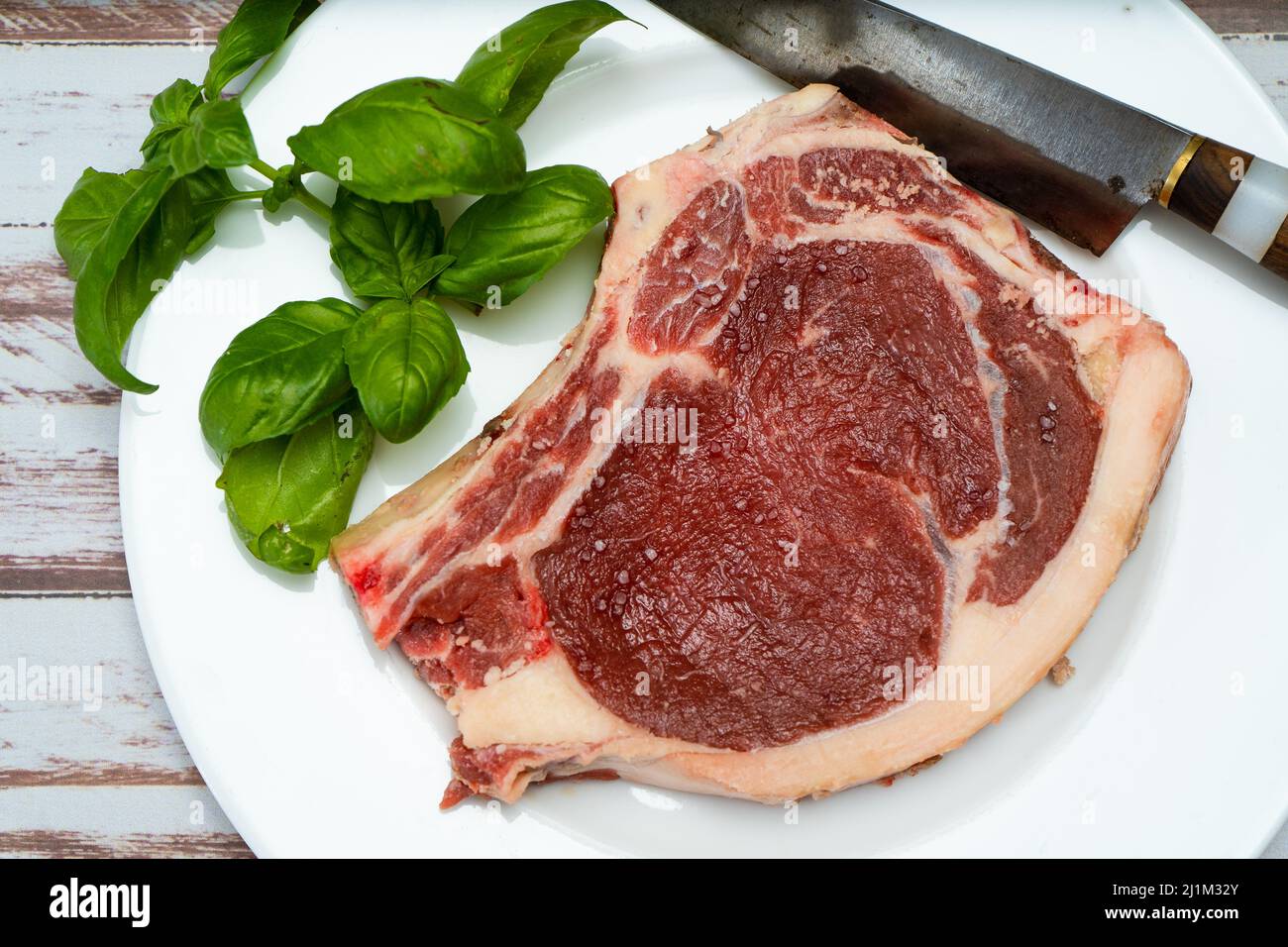 A bone-in steak or t-bone steak or porterhouse raw on a white plate on a wooden table with some green leaves. Top view. Horizontal orientation. Close- Stock Photo
