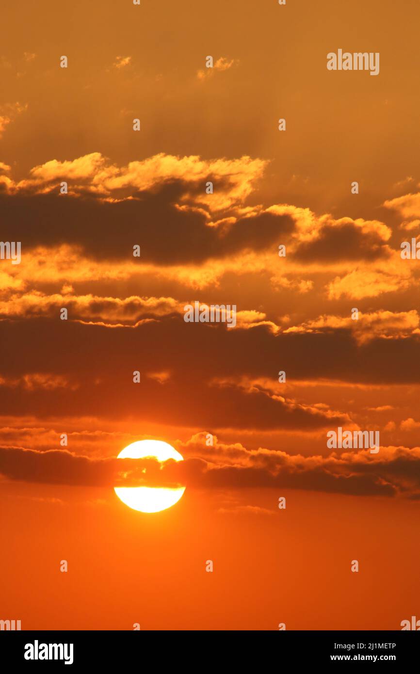 View of a colourful sunset on a March evening with the sun peeking out from behing sunset coloured cloud, some with golden yellow linings. Stock Photo